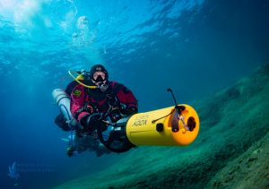 Nurek techniczny z wieloma butlami używa skutera podowdnego. Nurek w czerwonym suchym skafandrze i żółtym skuterem podwodnym