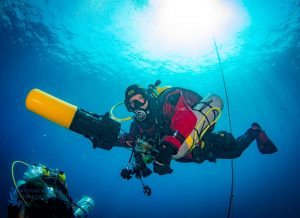 Nurek tchczniczny z wieloma butlami używa skutera podowdnego. Nurek w czerwonym suchym skafandrze i żółtym skuterem podwodnym